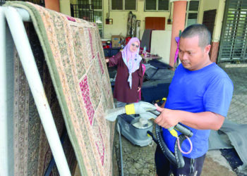 ZAMRI Muhamad menyedut habuk karpet menggunakan mesin sambil diperhatikan isterinya, Nurul Anis Ayob di rumah mereka di Bandar Utama, Gua Musang, Kelantan. – UTUSAN/AIMUNI TUAN LAH