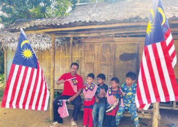 RAINA Anjang bersama kanak-kanak Orang Asli mengibar Jalur Gemilang di halaman rumahnya di Kampung Aring 5, Gua Musang, Kelantan.-UTUSAN/AIMUNI TUAN LAH