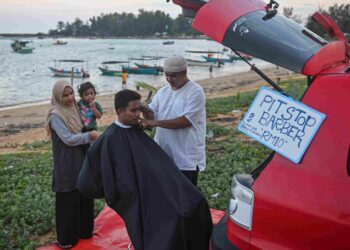 NUR Imran Hasmady menggunting rambut seorang pelanggannya, Muhammad Syakir Abdul Latif di kawasan rekreasi, Pantai Kekabu, Marang, Terengganu, semalam. – UTUSAN/PUQTRA HAIRRY ROSLI