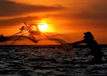 Seorang nelayan menjala berlatar belakang matahari terbenam di lautan.