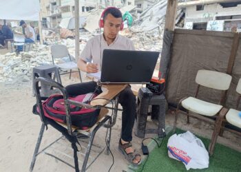 ABDALLAH Abuajwa mengikuti ujian kelayakan pengajian peringkat Doktor Falsafah (PhD) USIM secara dalam talian di bawah khemah di kawasan terbuka dengan berlatar belakang kawasan kediaman yang musnah dibedil tentera Israel di Gaza, Palestin.-GAMBAR/IHSAN USIM.