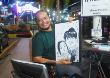 SHAHRIL Anuar Mohd. menunjukkan hasil karikatur wajah dua pelanggannya di Dataran Quayside, Banda Hilir, Melaka. 
– UTUSAN/AMRAN MULUP