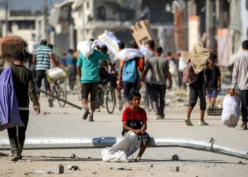 SEORANG kanak-kanak duduk di atas tiang lampu jalan yang tumbang ketika orang ramai berjalan membawa bungkusan bantuan kemanusiaan daripada pusat agihan GHF di koridor Netzarim di Nuseirat, tengah Gaza.-AFP