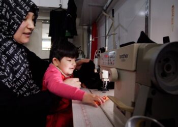 KALBINUR menjahit pakaian sambil memangku anaknya, Merziye. - BBC