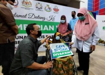 HASNI Mohammad (dua kiri) menyantuni warga emas, Sadinah Karman yang menerima vaksin di PPV Benut bersama-sama Dr. Surinah Asan (kanan) di Dewan Serbaguna Benut, di Pontian, Johor.