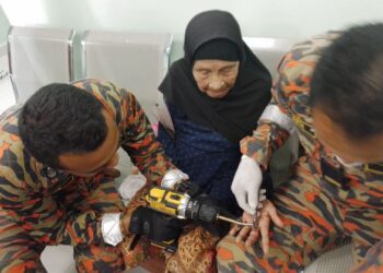 DUA anggota bomba sedang menanggalkan cincin emas Munah Tawang yang tersangkut di jari tengah tangan kirinya di Klinik Kesihatan Chegar Perah di Lipis, Pahang. - FOTO/IHSAN BALAI BOMBA KUALA LIPIS