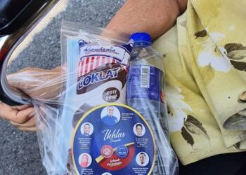 GAMBAR tular memaparkan seorang penerima vaksin menerima sumbangan yang tertera logo UMNO dan BN ketika mendapatkan suntikan di PPV, Simpang Renggam, Johor.